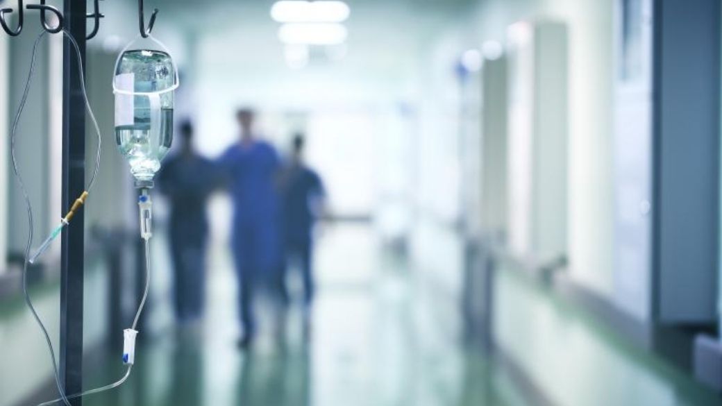In the foreground, an IV bag hangs on a metal IV pole. In the background, three healthcare workers are seen out of focus at the end of a hallway.